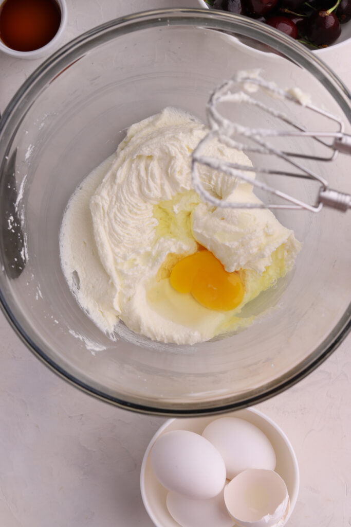 An egg being mixed into cake batter and a bowl of cherries. 