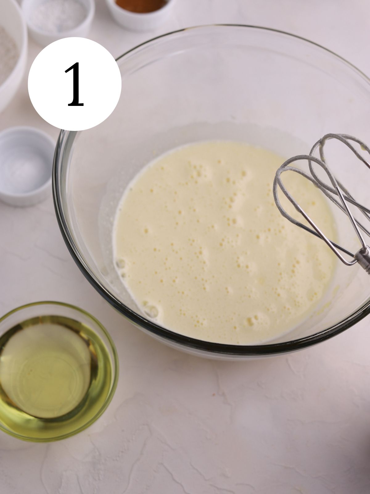 A glass mixing bowl with eggs and sugar combined by an electric mixer.