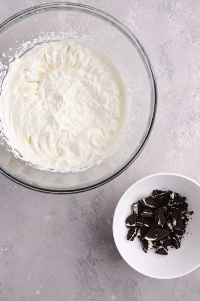 A bowl of whipped cream and a small bowl with crushed Oreo cookies.