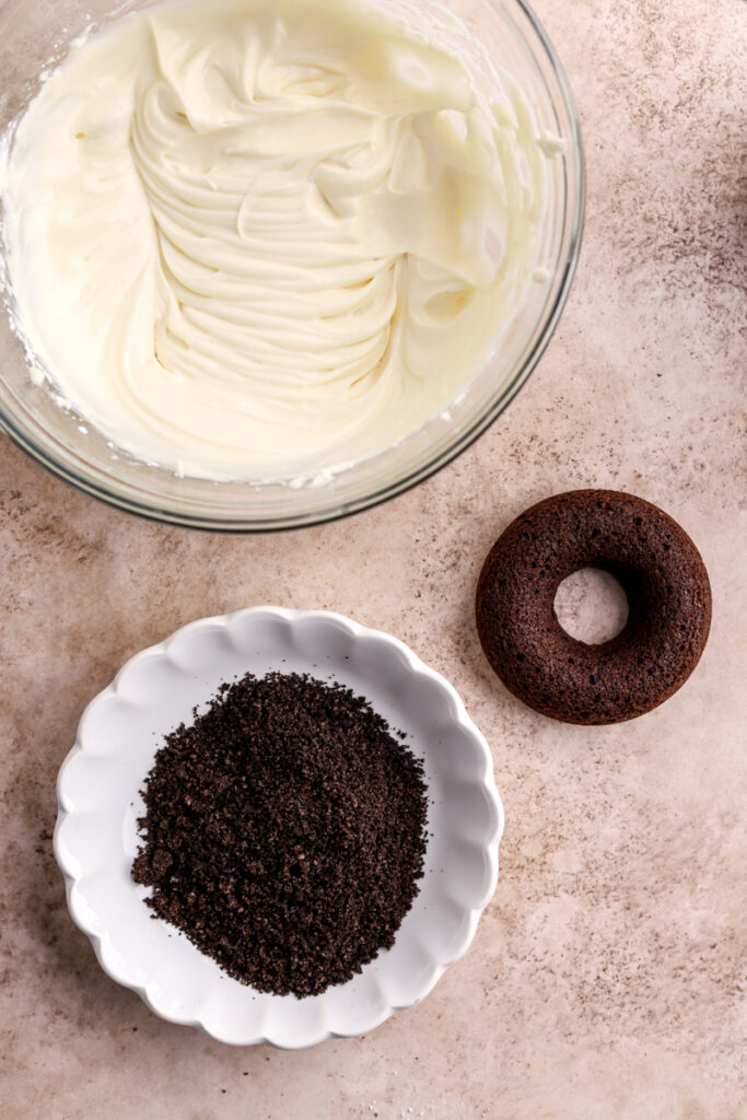 A glass bowl with cream cheese icing, a chocolate donut, and a bowl of Oreo crumbs.
