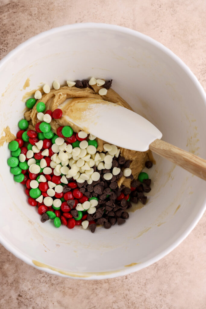 Red and green M&Ms, some chocolate chips, cream cheese chips and a spatula in a white bowl.