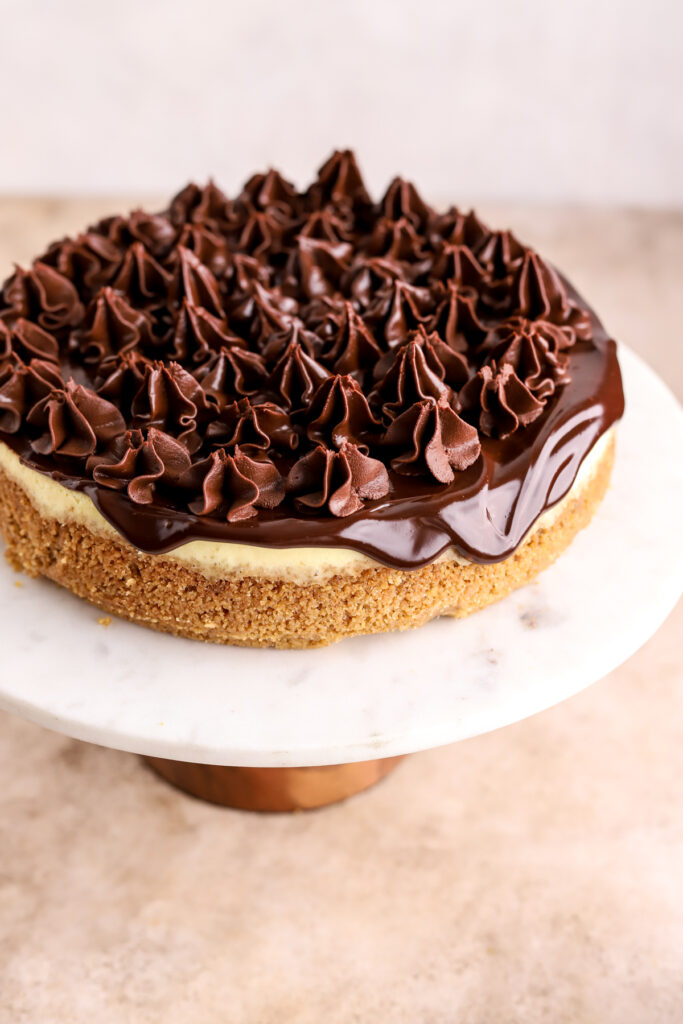 A cheesecake topped with chocolate ganache and chopped Toblerone on a cake stand.