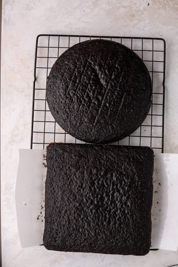 A black cooling rack with a round and a square chocolate cake on it. 