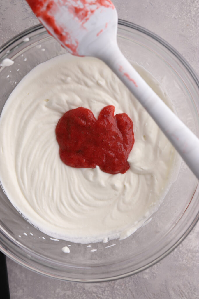 A bowl of whipped cream with pureed strawberries on top and a spatula.