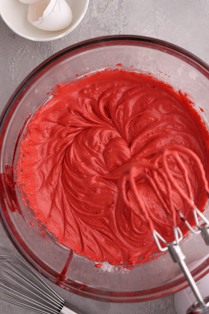A glass mixing bowl with red velvet batter in it and a mixer.