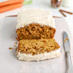 A loaf cake with frosting, a knife, and some carrots and cinnamon sticks.