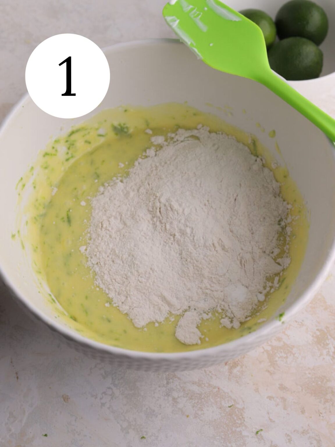 A white bowl with cake batter, flour and a spatula and a bowl of limes.