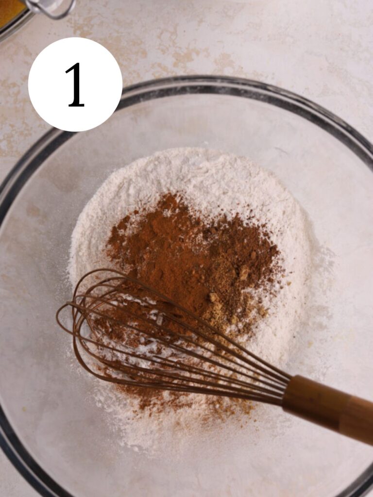 A glass mixing bowl with spices, flour and a whisk. 