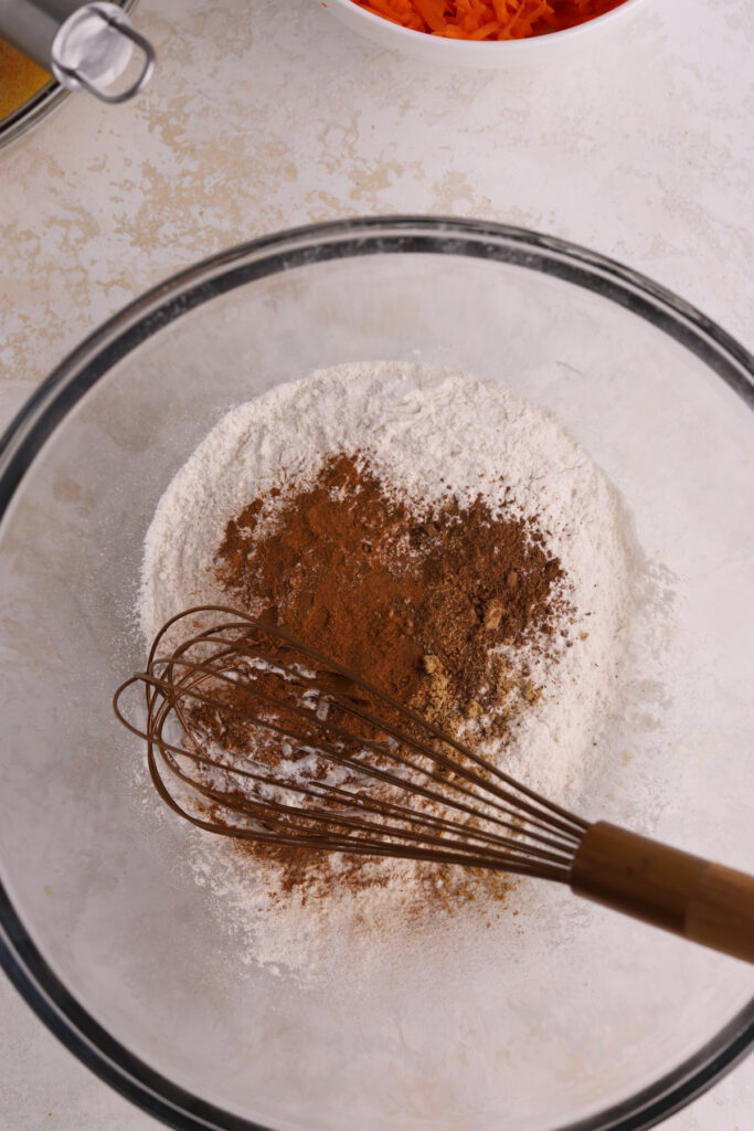 A glass mixing bowl with spices, flour and a whisk. 