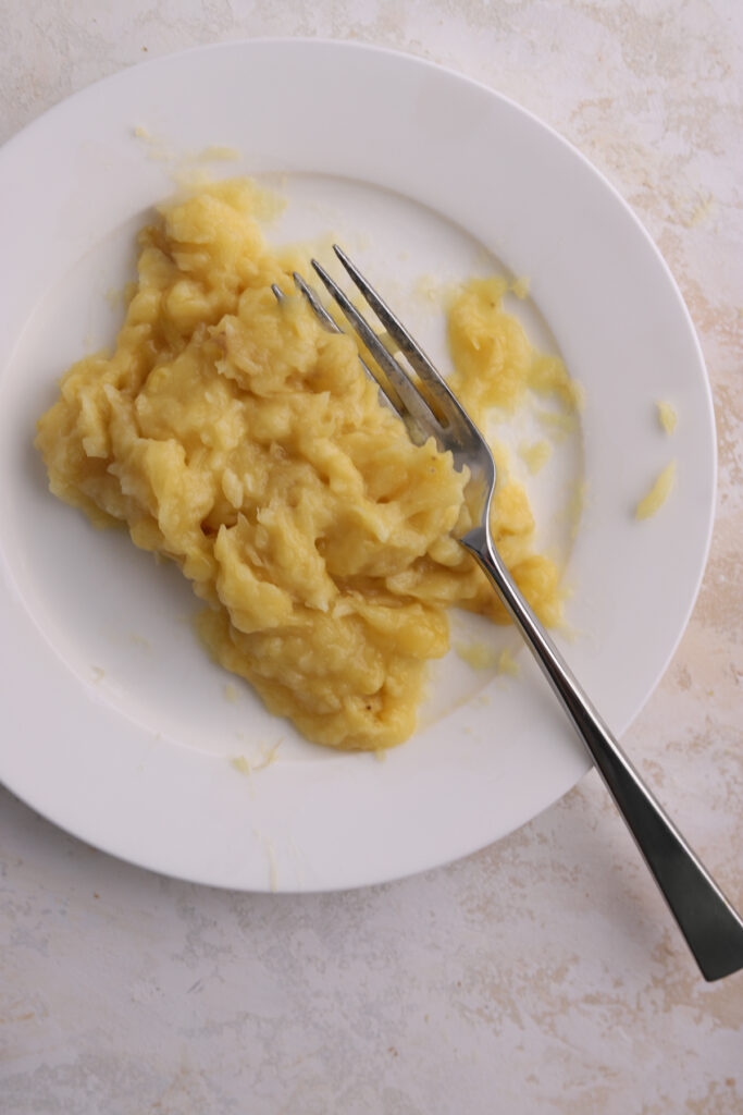 Banana being mashed with a fork on a plate. 