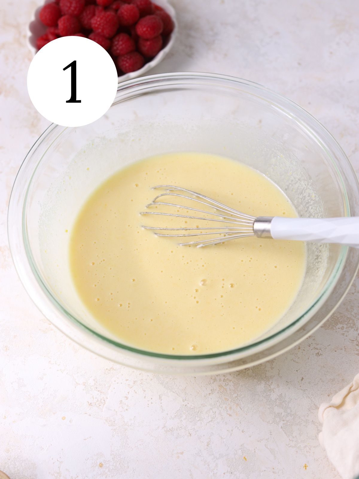 Wet ingredients in a mixing bowl with a whisk and a bowl of raspberries.
