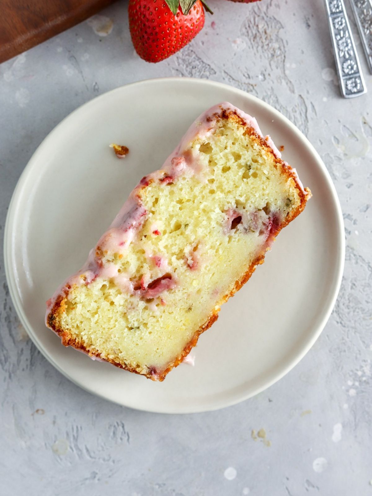 A slice of strawberry loaf cake aith glaze on top on a plate next to some fresh strawberries. 
