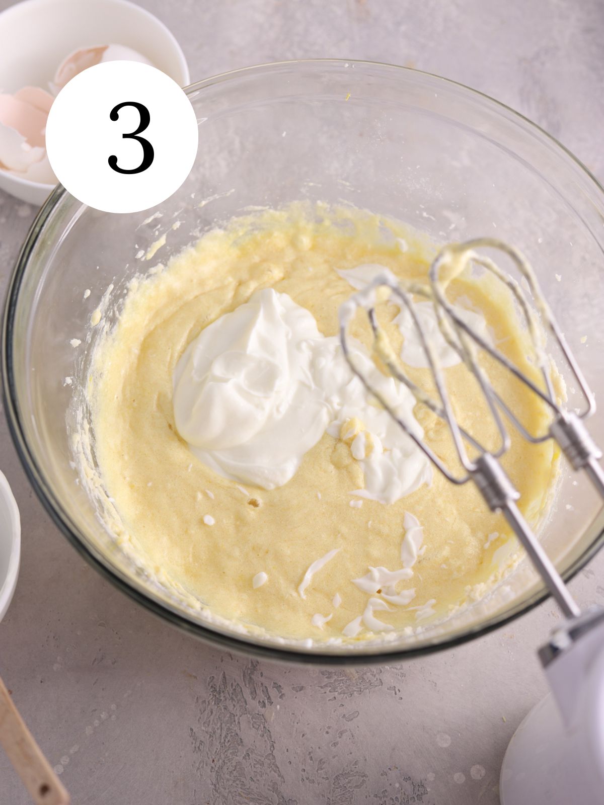 Yogurt added to cake batter in a glass mixing bowl next to an electric mixer. 