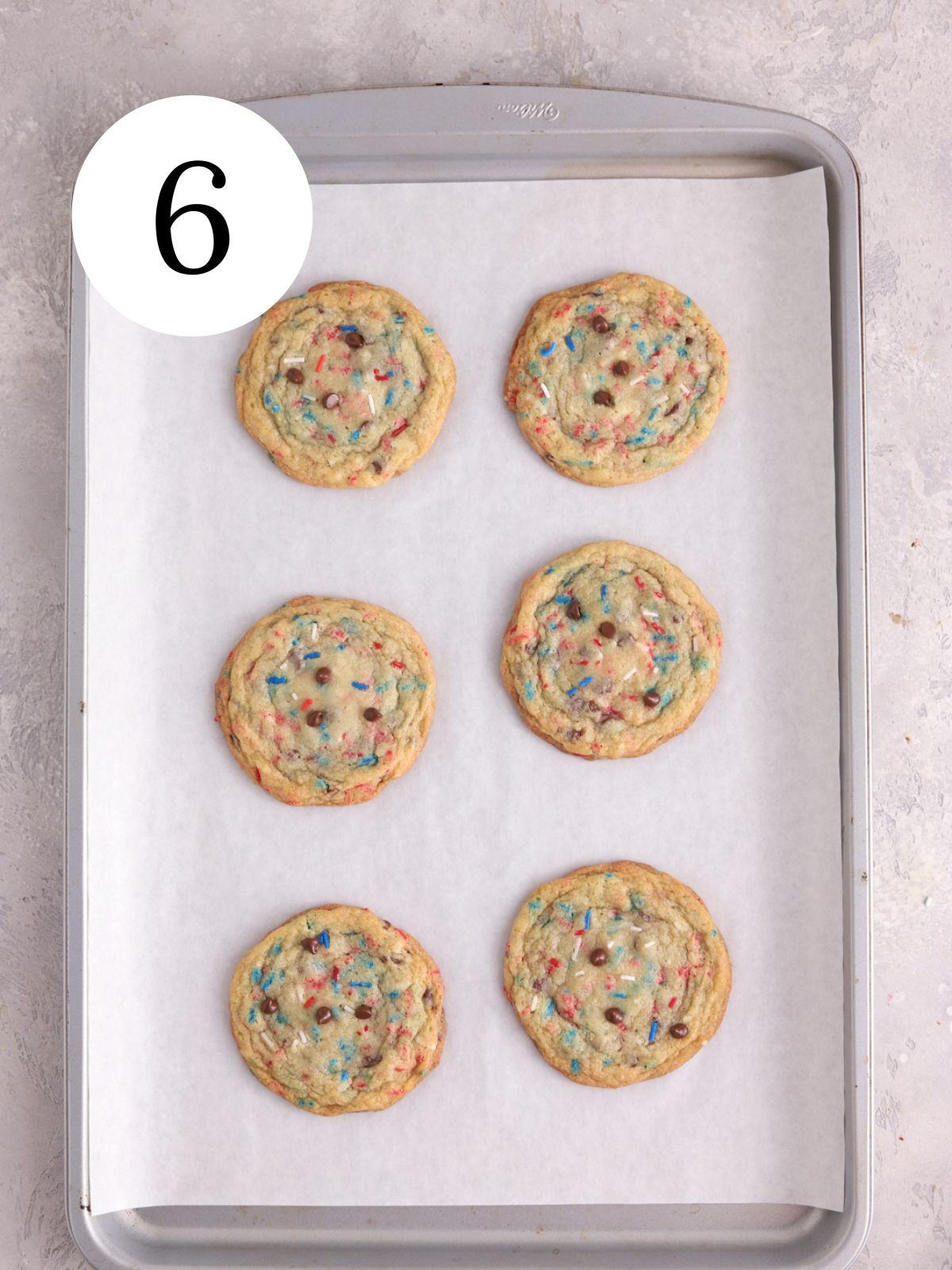 Six chocolate chip sprinkle cookies on a baking sheet lined with parchment paper.