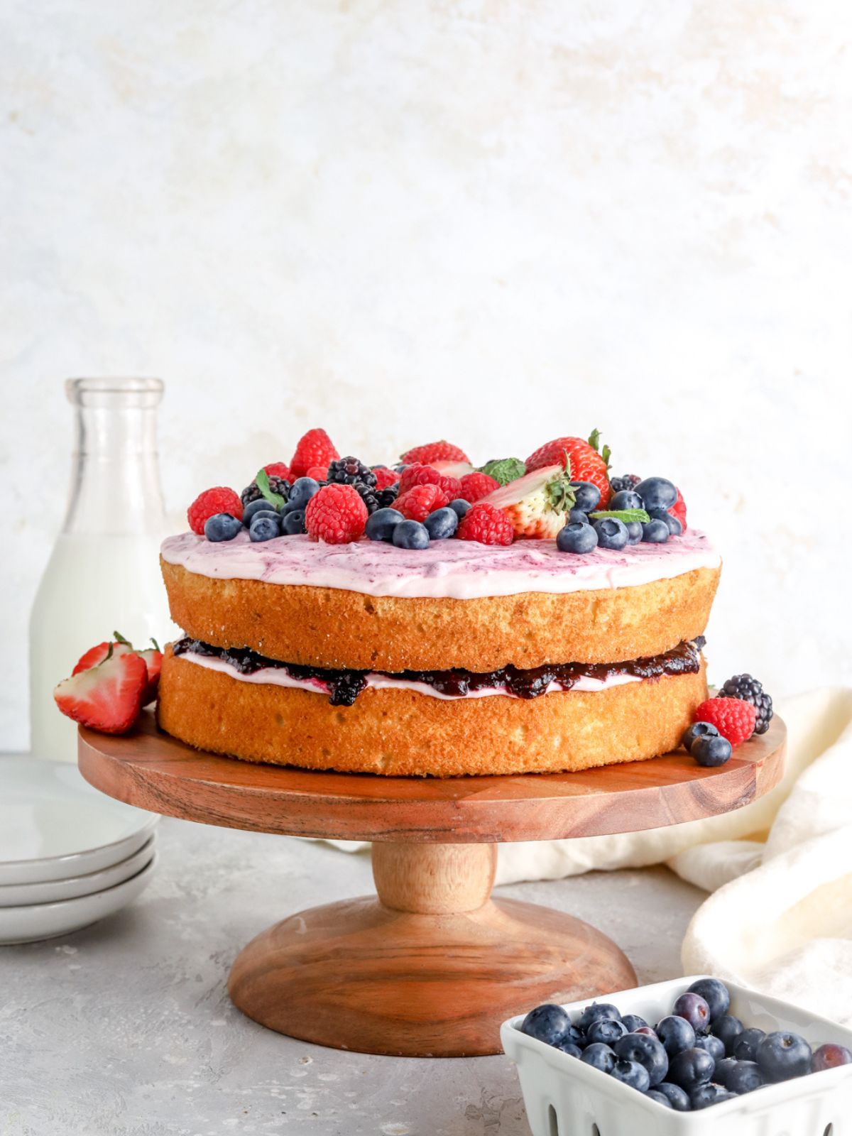 A two layer cake with jam and frosting and berries on a cake stand.