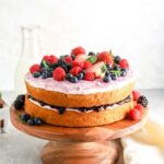 A two layer cake on a cake stand with jam and frosting in the middle and berries on top.