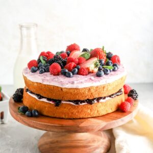 A two layer cake on a cake stand with jam and frosting in the middle and berries on top.