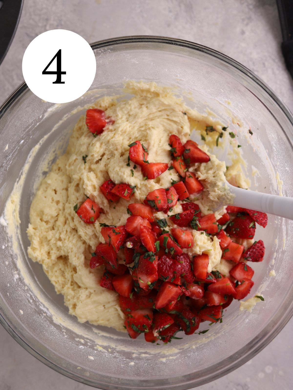 Cake batter in a glass bowl with fresh strawberries and chopped tarragon being stirred in. 