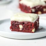 A frosted red velvet brownie with white chocolate on a white plate.