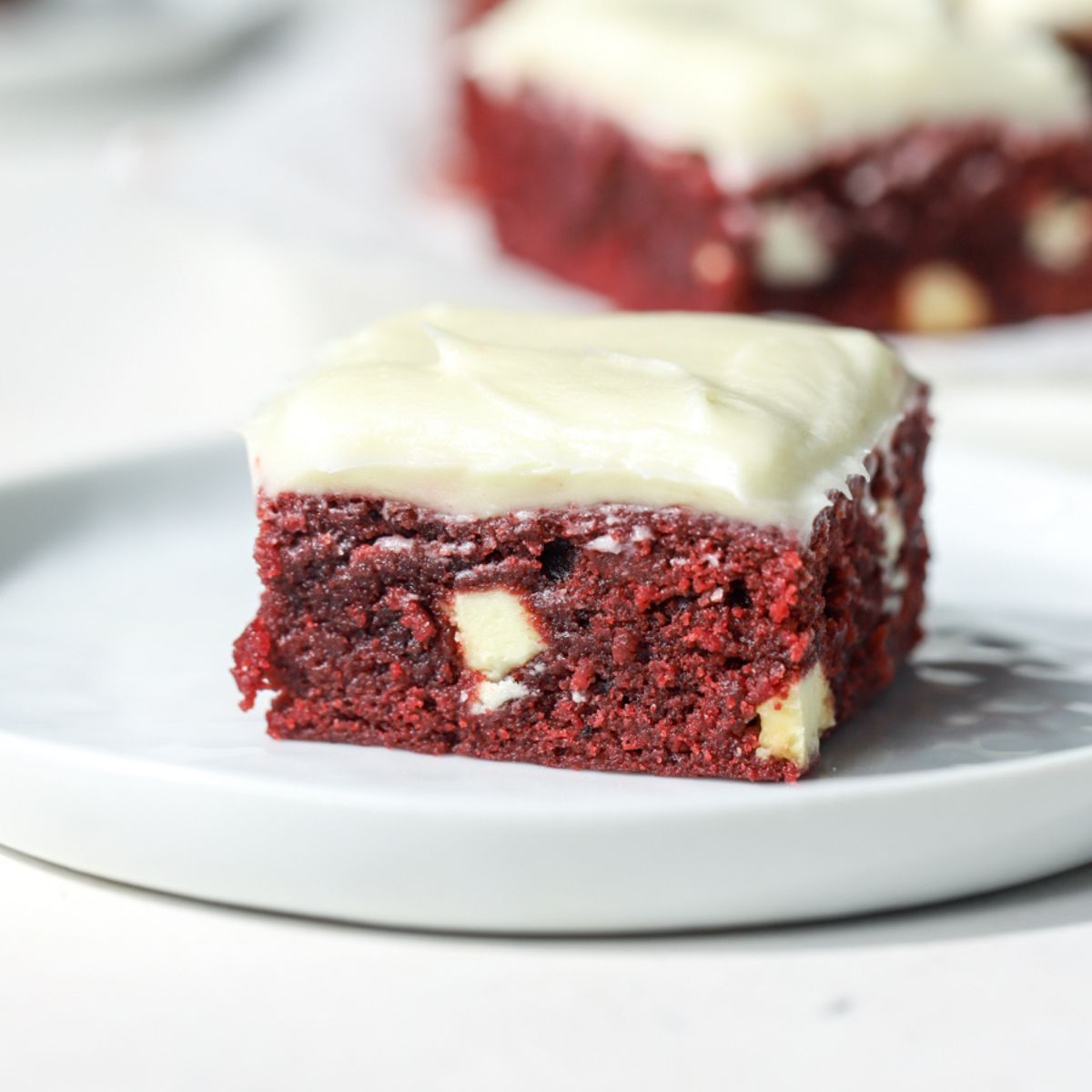 A frosted red velvet brownie with white chocolate on a white plate.