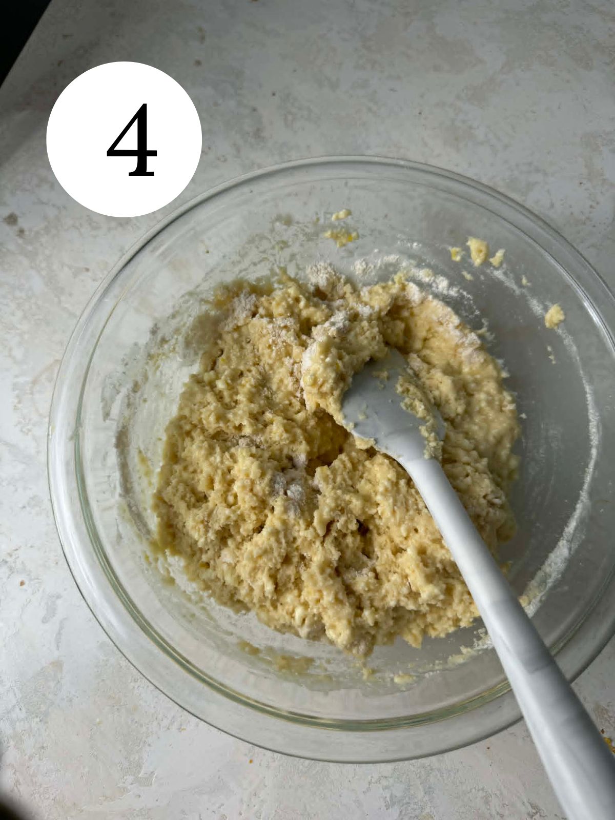Flour being stirred into ricotta batter with a spatula in a glass bowl.