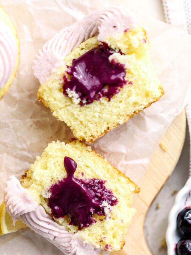 A frosted cupcake cut in half with a purple blueberry filling and a bowl of blueberries.
