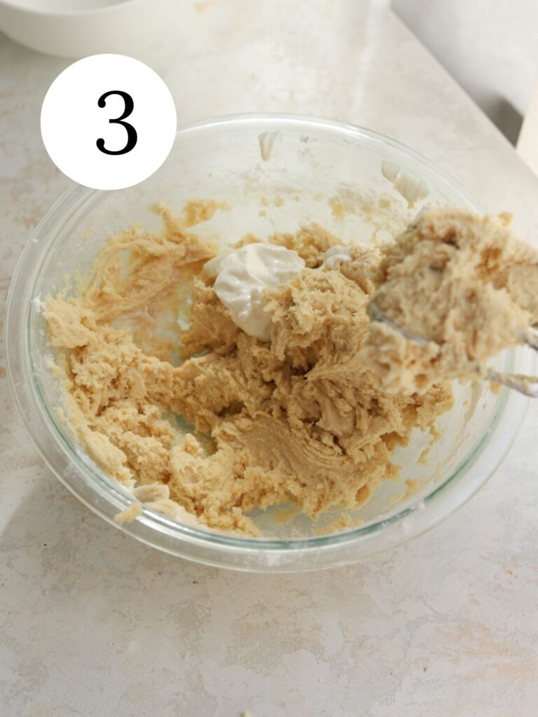 Adding sour cream to cookie dough in a glass bowl. 