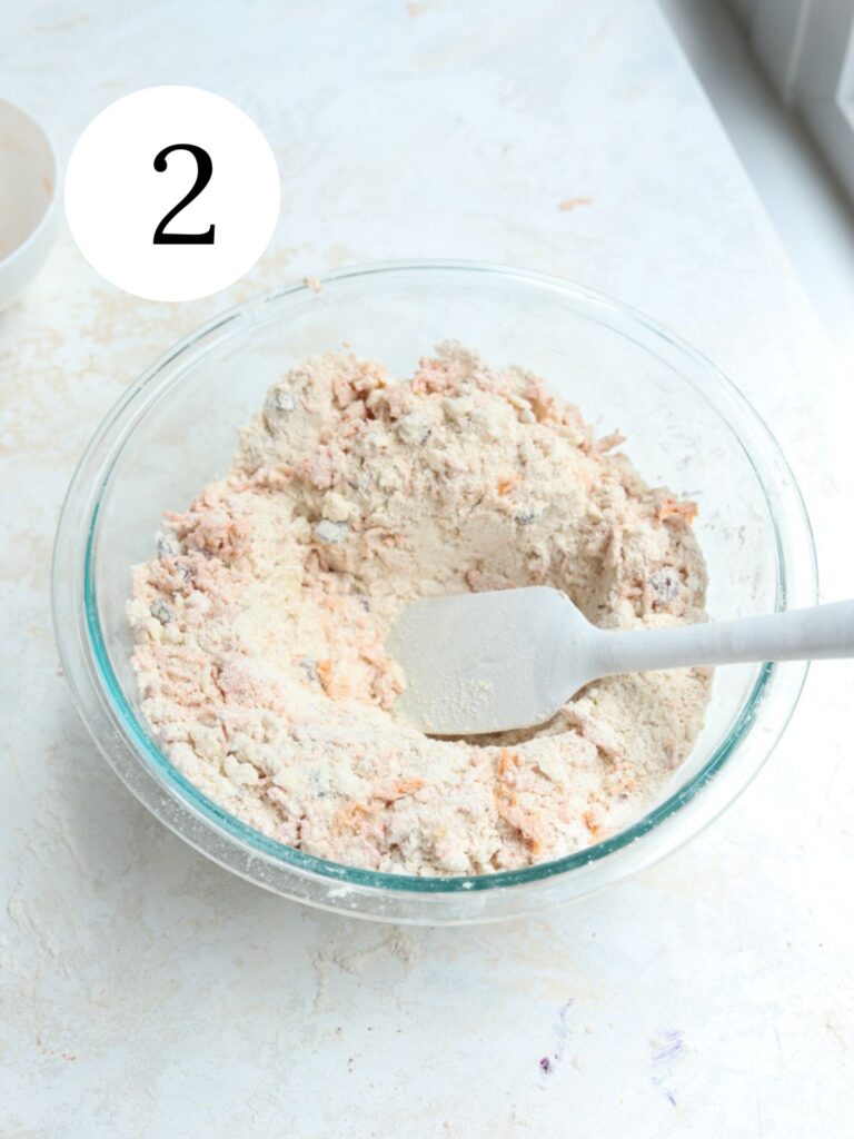 A glass mixing bowl with flour and carrots and a spatula in it. 