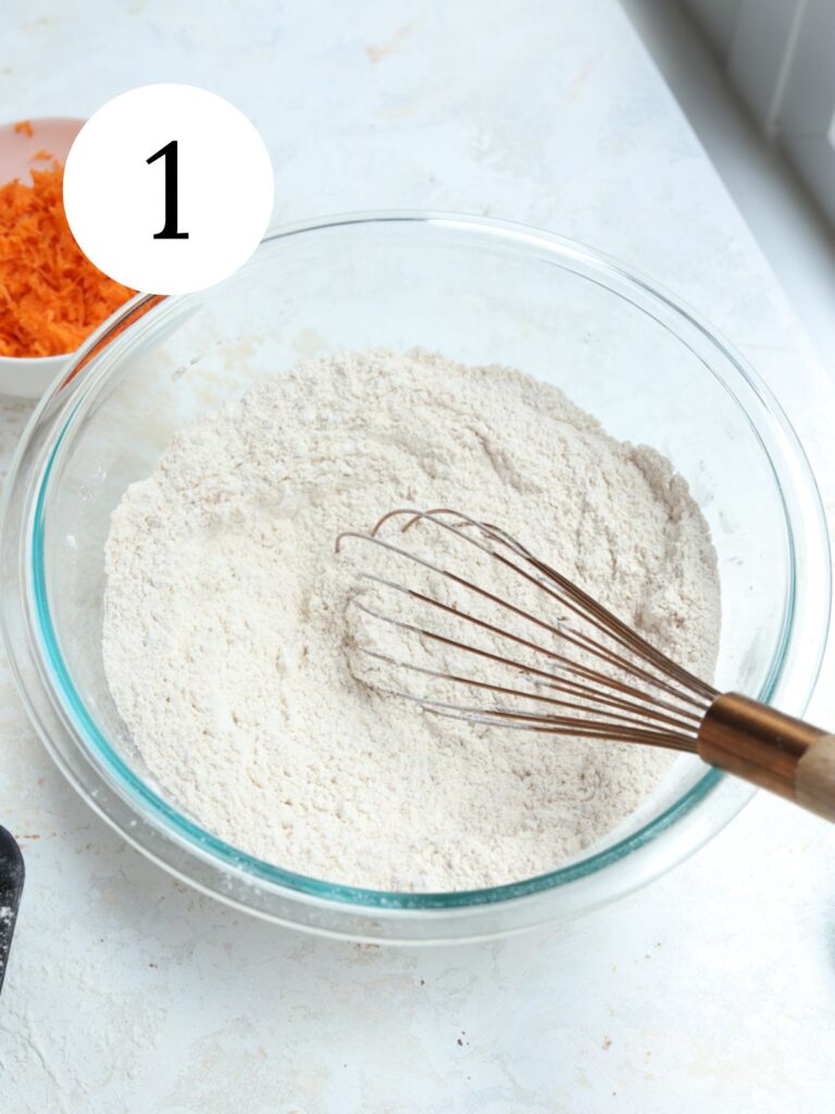 A glass bowl with dry ingredients and a whisk next to a bowl of shredded carrots. 