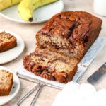A loaf of Nutella banana bread being sliced on a rack next to some bananas.
