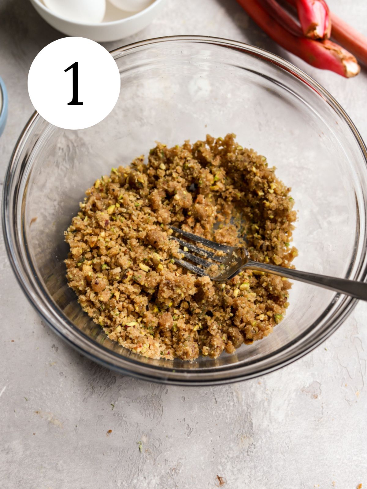 Crumble topping in a glass bowl with a fork next to some eggs and rhubarb.