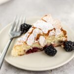 A piece of cake with blackberries topped with almonds and powdered sugar on a plate.