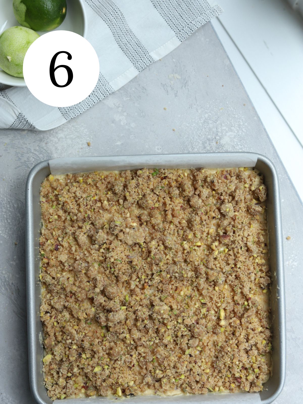 Cake with crumble topping in a square metal bowl next to some limes.