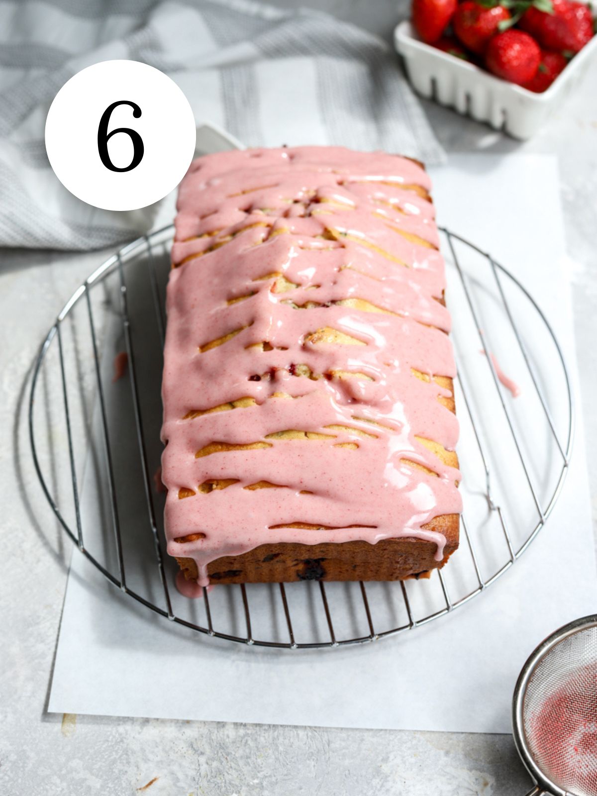 A loaf cake with strawberry icing on a wire rack and a bowl of strawberries.