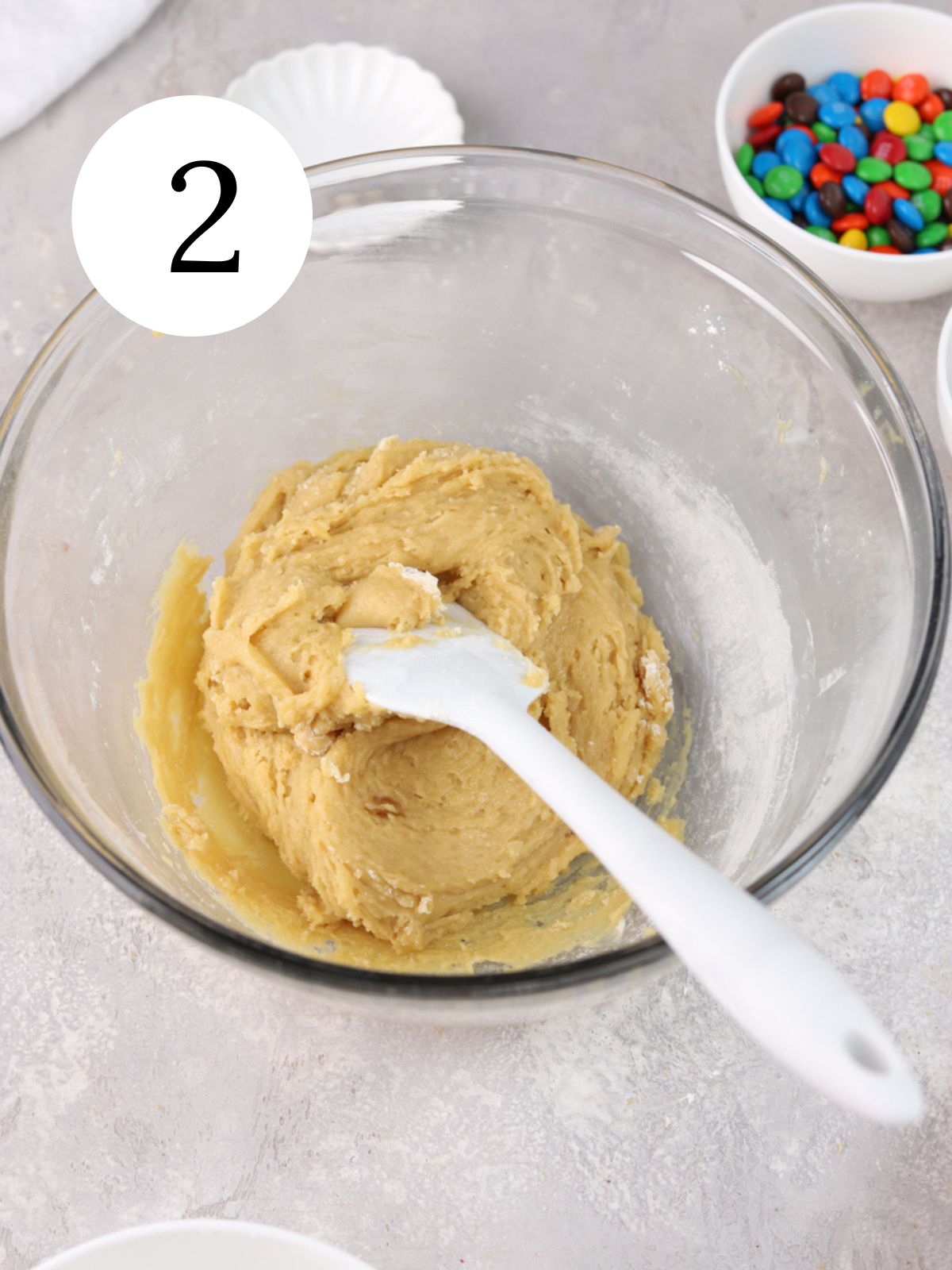 A mixing bowl with a spatula and cookie dough and a white bowl with M&M candy.