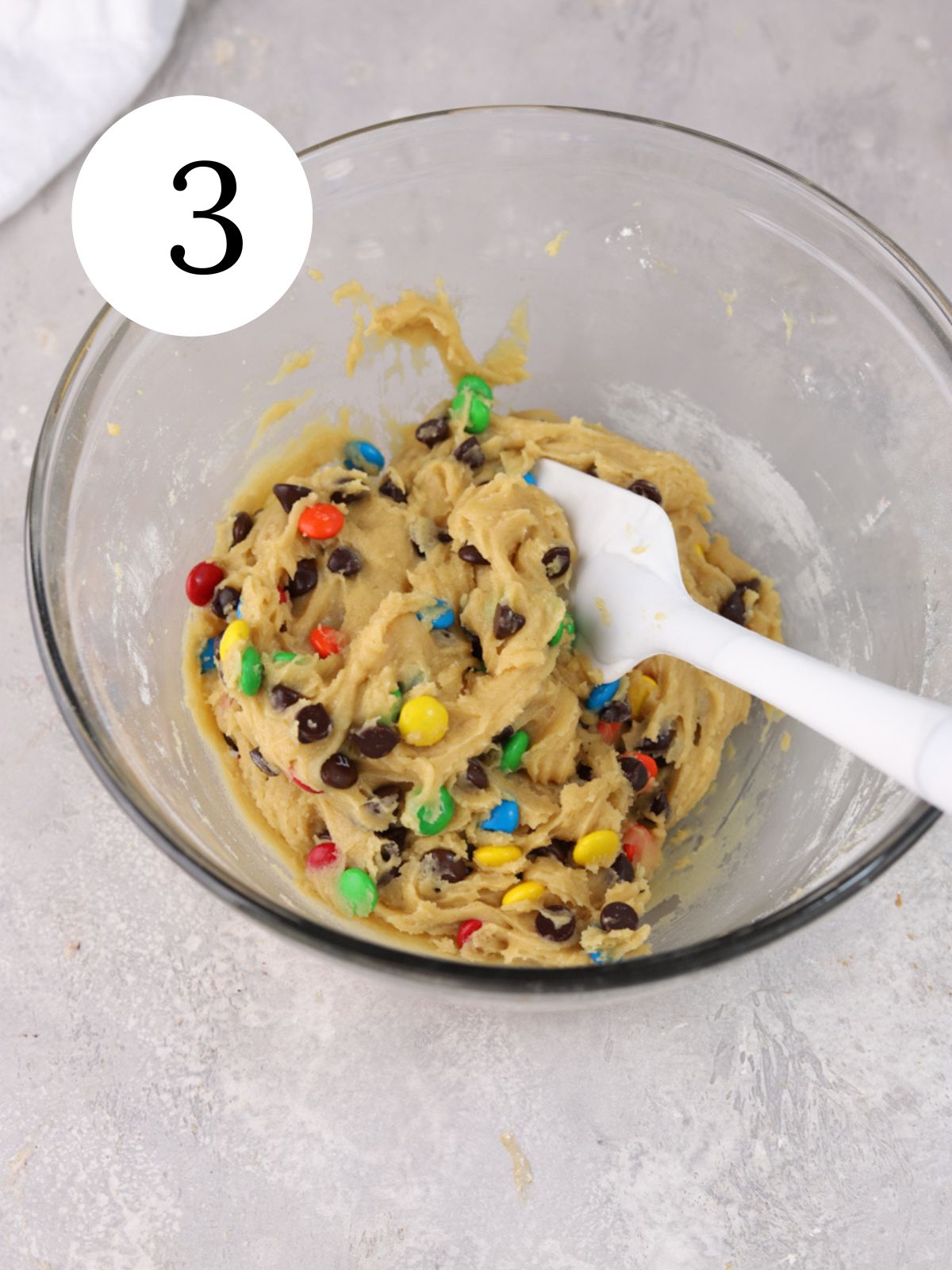 A glass mixing bowl with chocolate chip M&M cookie dough and a spatula.