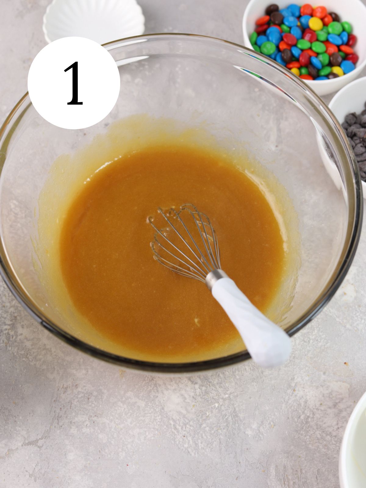 A glass mixing bowl with melted butter and sugars and a whisk.