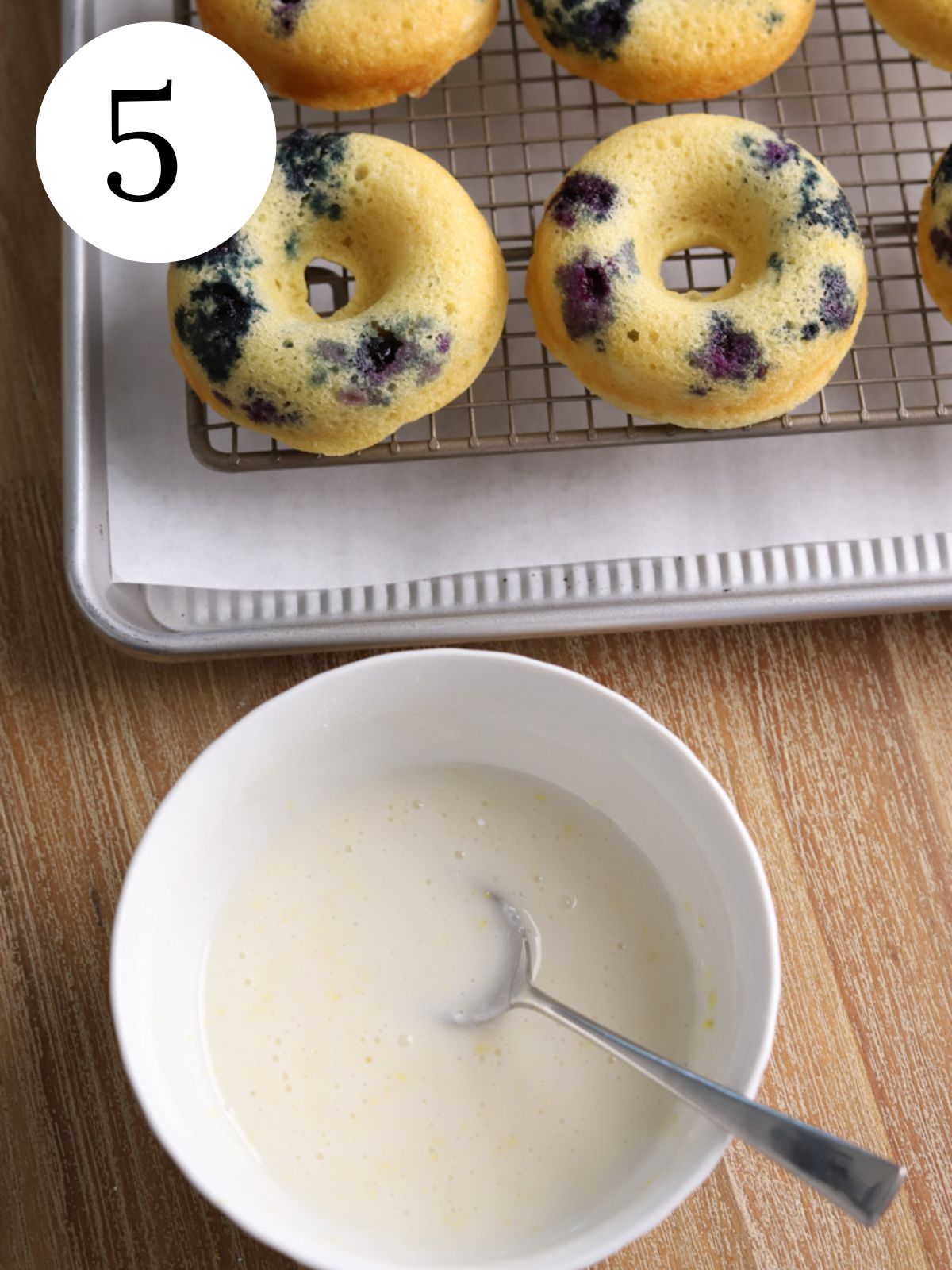 A bowl with icing and a spoon and some blueberry donuts on a rack. 
