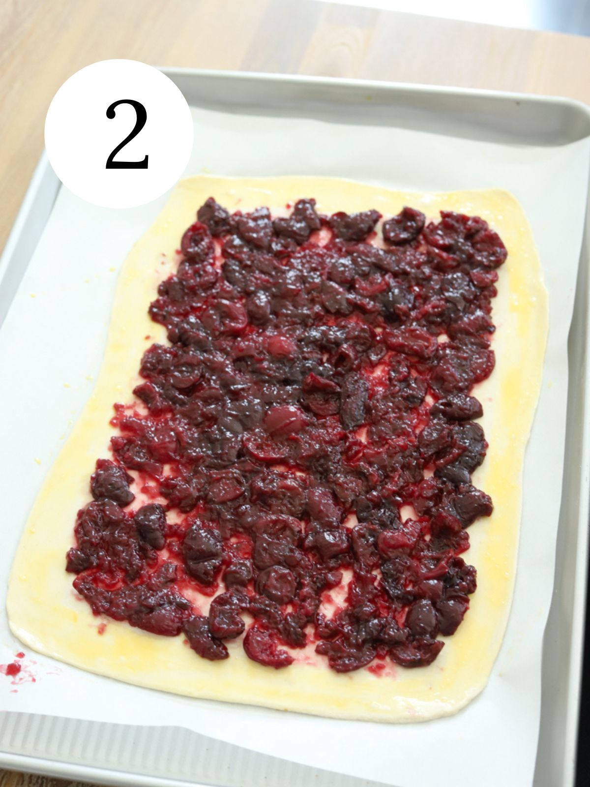 A rectangular pie crust on a baking sheet with cherry sauce spread on it.
