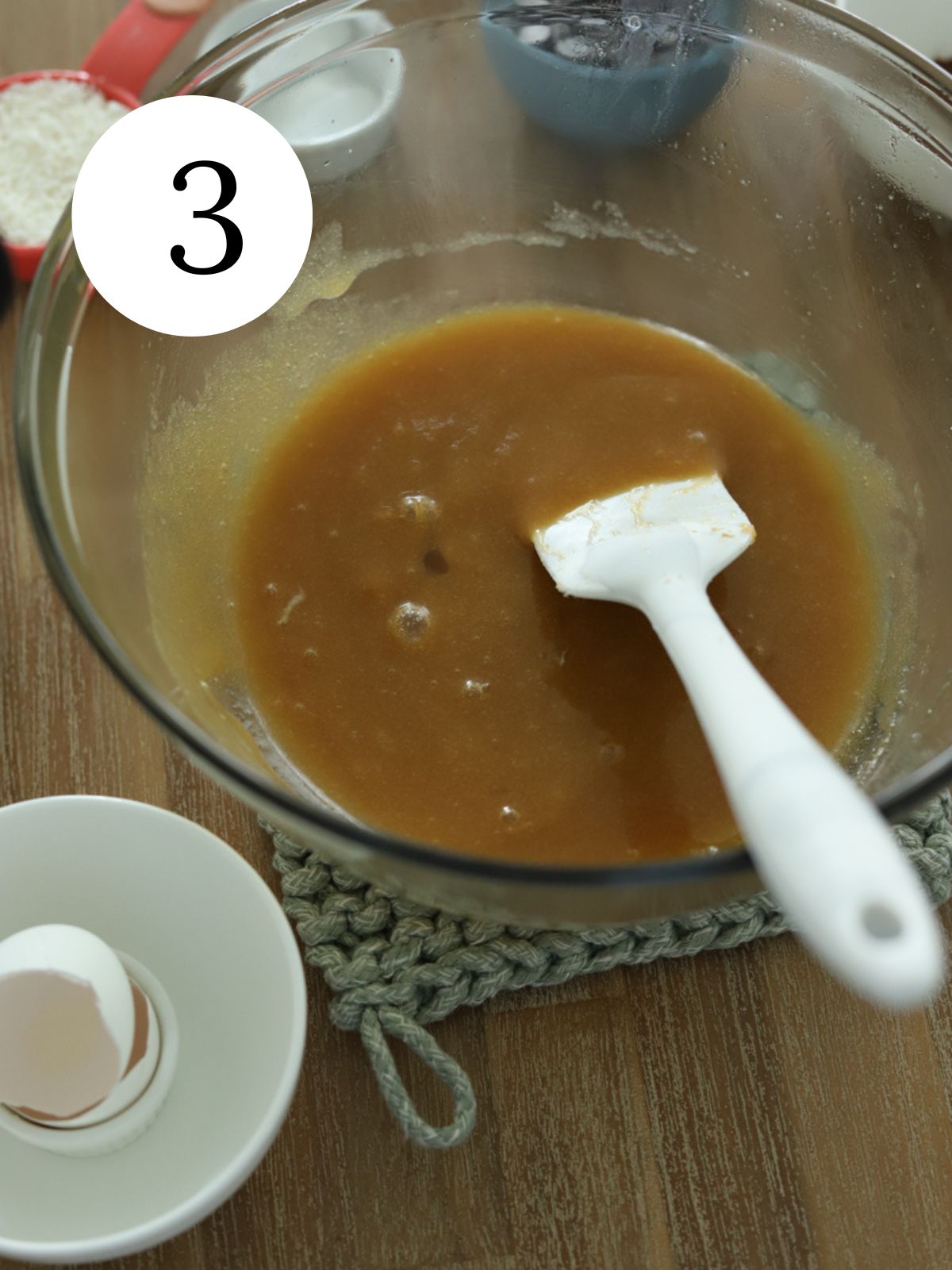 A glass bowl with dough being stirred with a spatula and a bowl with an egg shell. 