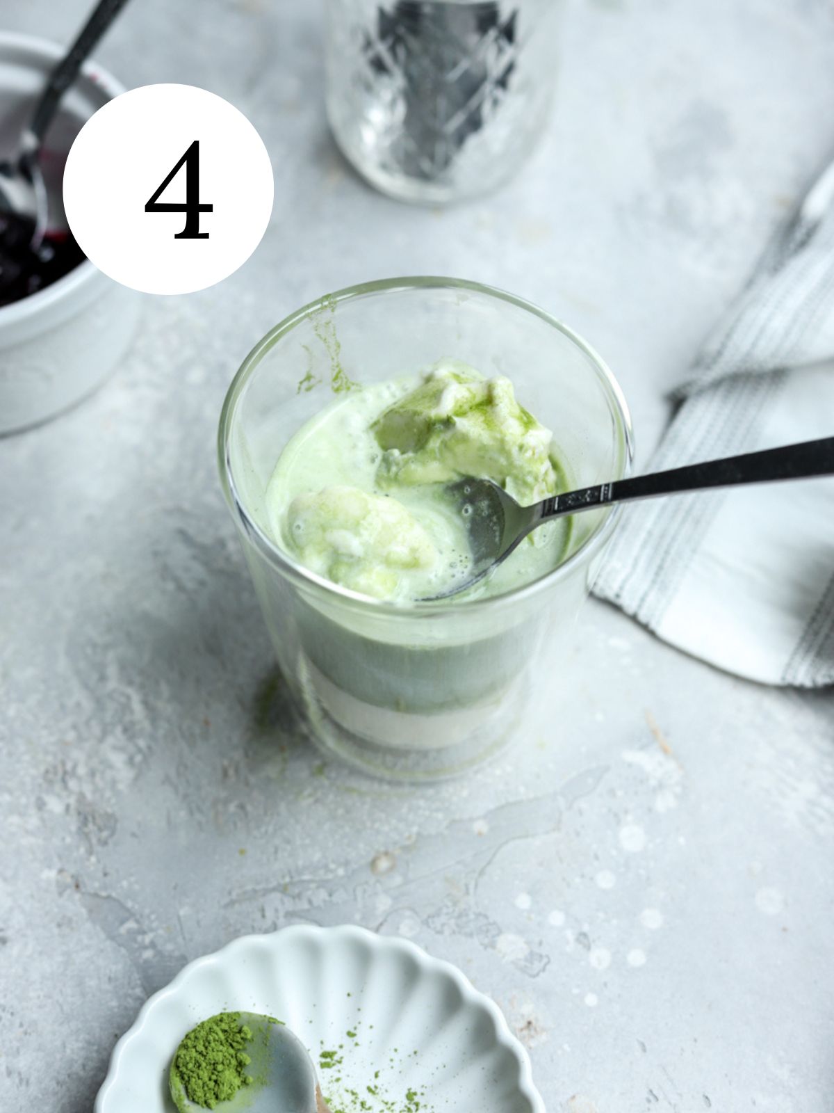 A glass with ice cream and matcha with a spoon in it and a dish with matcha powder.