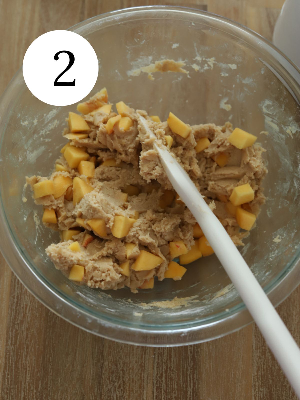 Chopped peaches stirred into cookie dough with a spatula in a glass bowl. 