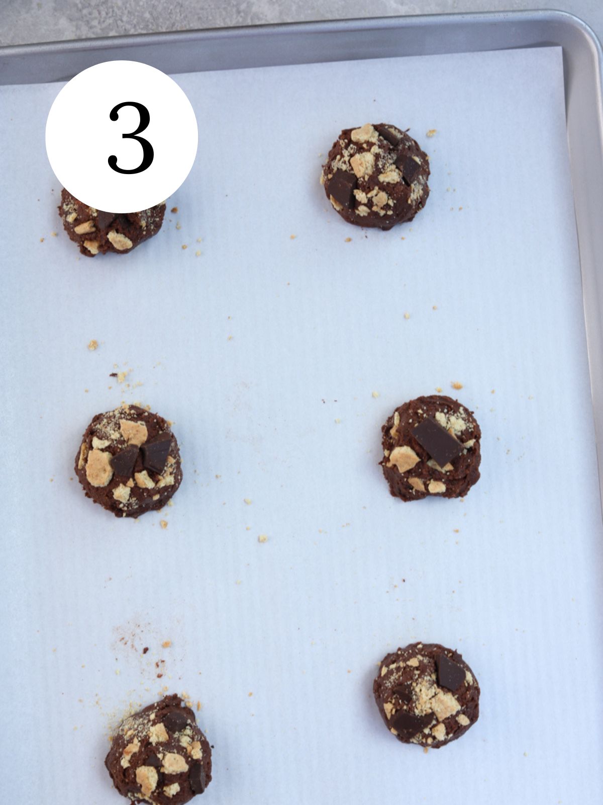 Chocolate cookie dough balls topped with graham cracker crumbs and chocolate on a baking sheet.