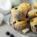 Blueberry muffins stacked on each other on a wire rack next to some blueberries.