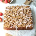 A square cake with crumble and icing on top next to a bowl of strawberries.