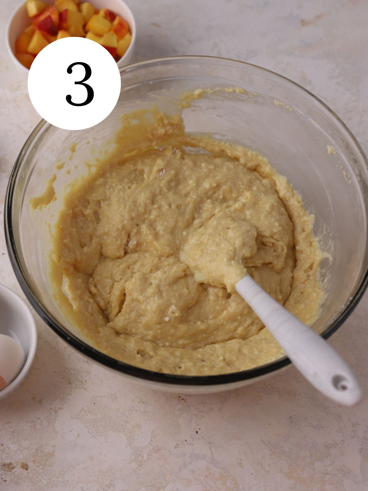 A mixing bowl with cake batter and a spatula next to a bowl of peaches.