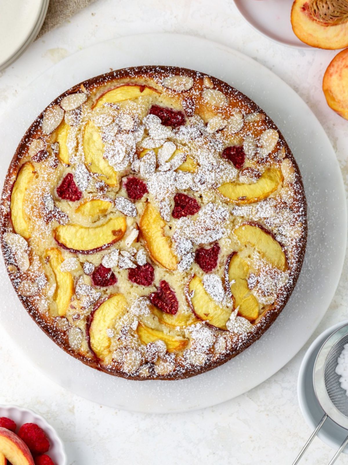 A cake topped with peaches and raspberries and dusted with powdered sugar on a white marble tray.