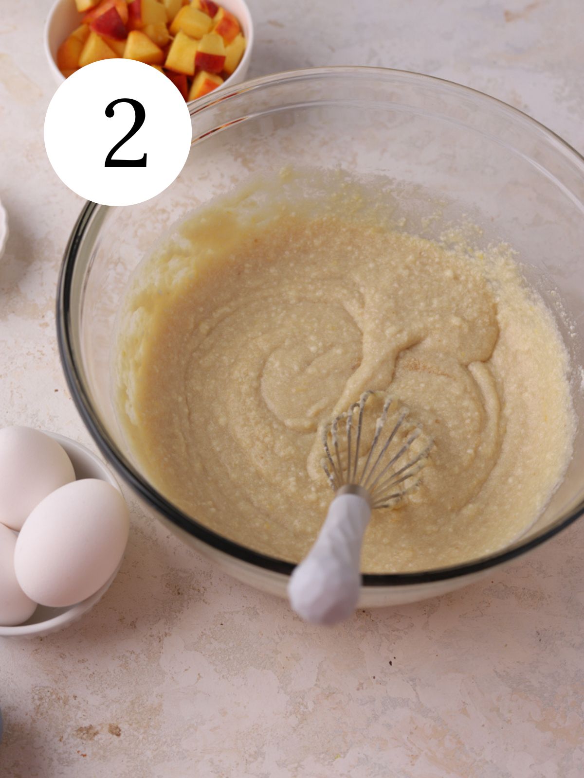 A bowl with a whisk and cake batter in it next to some eggs and chopped peaches.
