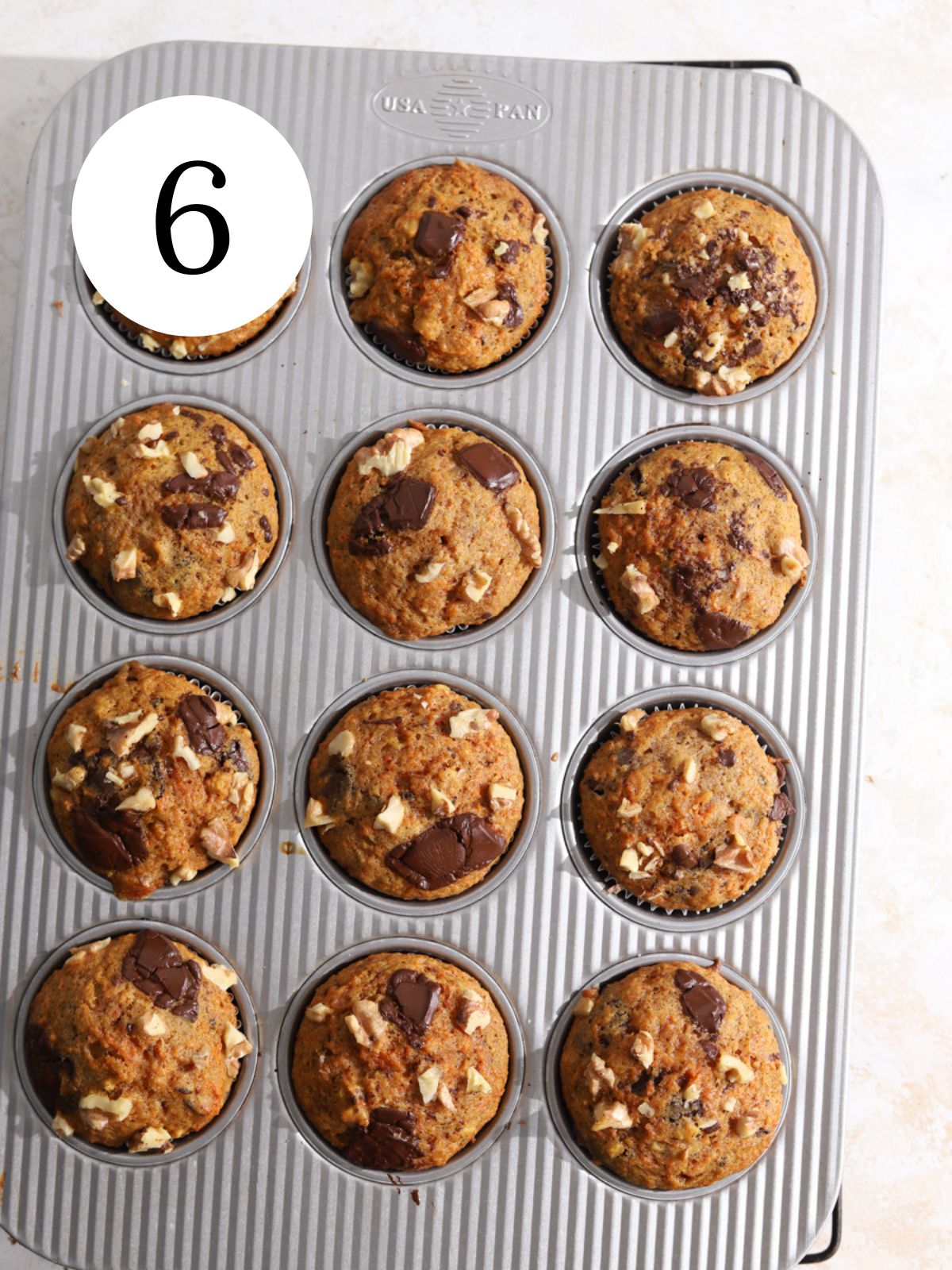 A muffin pan with a batch of carrot muffins topped with chocolate and walnuts.