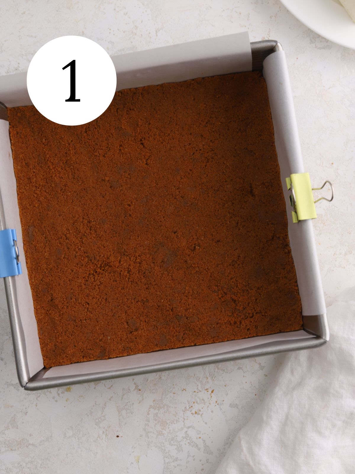 A biscoff cookie crust in a square pan lined with parchment paper.